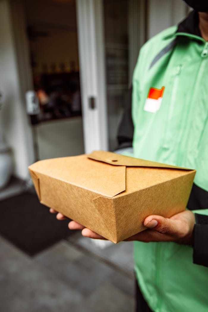 Close-up of a delivery person holding a brown paper food box near a restaurant.